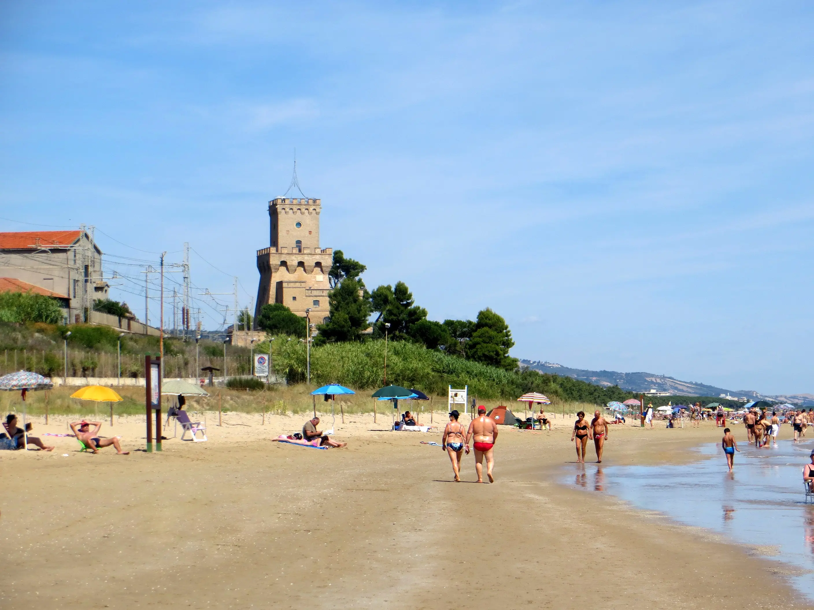 Bagnanti passeggiano sulla battigia con la Torre del Cerrano sullo sfondo, in piena estate.